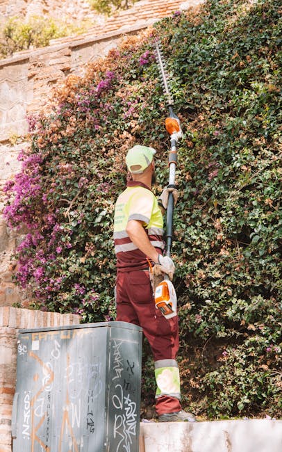 A gardener wearing a light-colored cap, high-visibility vest, and maroon work trousers is trimming a large, dense hedge with an extendable pruning tool. The hedge, composed of small, green, and pink f